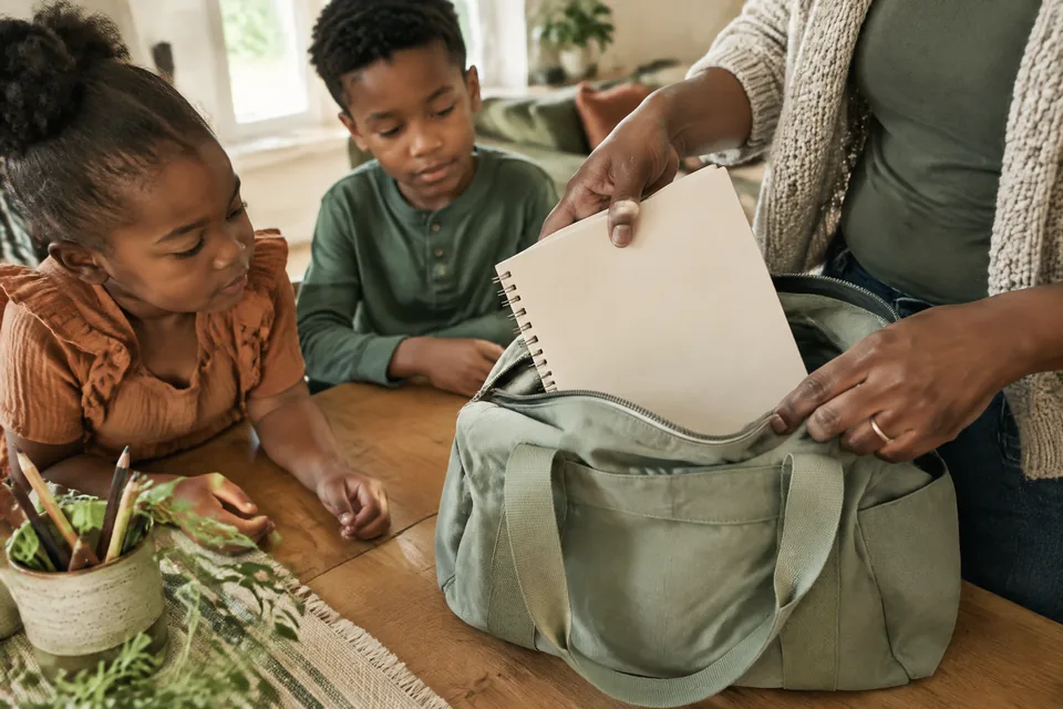A parent's hands sliding a blank drawing notebook into an open sage cotton busy bag on an oak table.