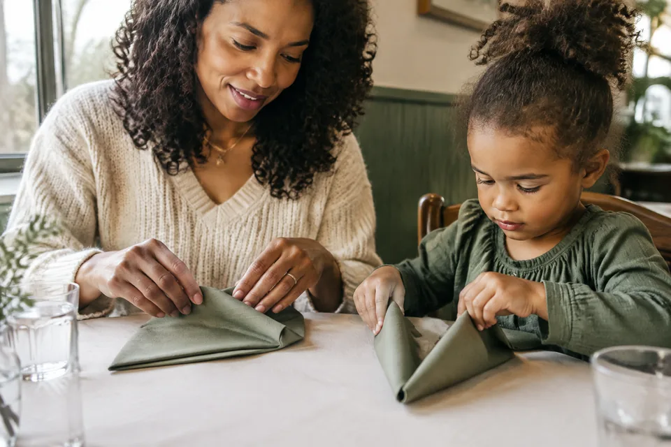 A parent and child making playful shapes with large folded paper napkins on a clean restaurant table, glasses placed away from hands.