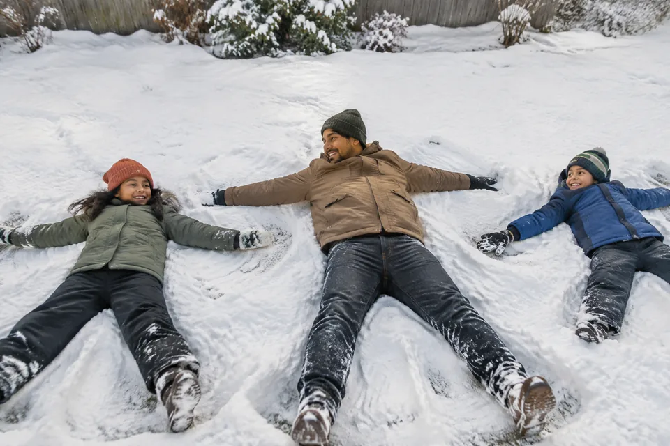 Parent and two school-age children making broad snow angels on a safe flat snowy backyard lawn, winter coats and mittens.